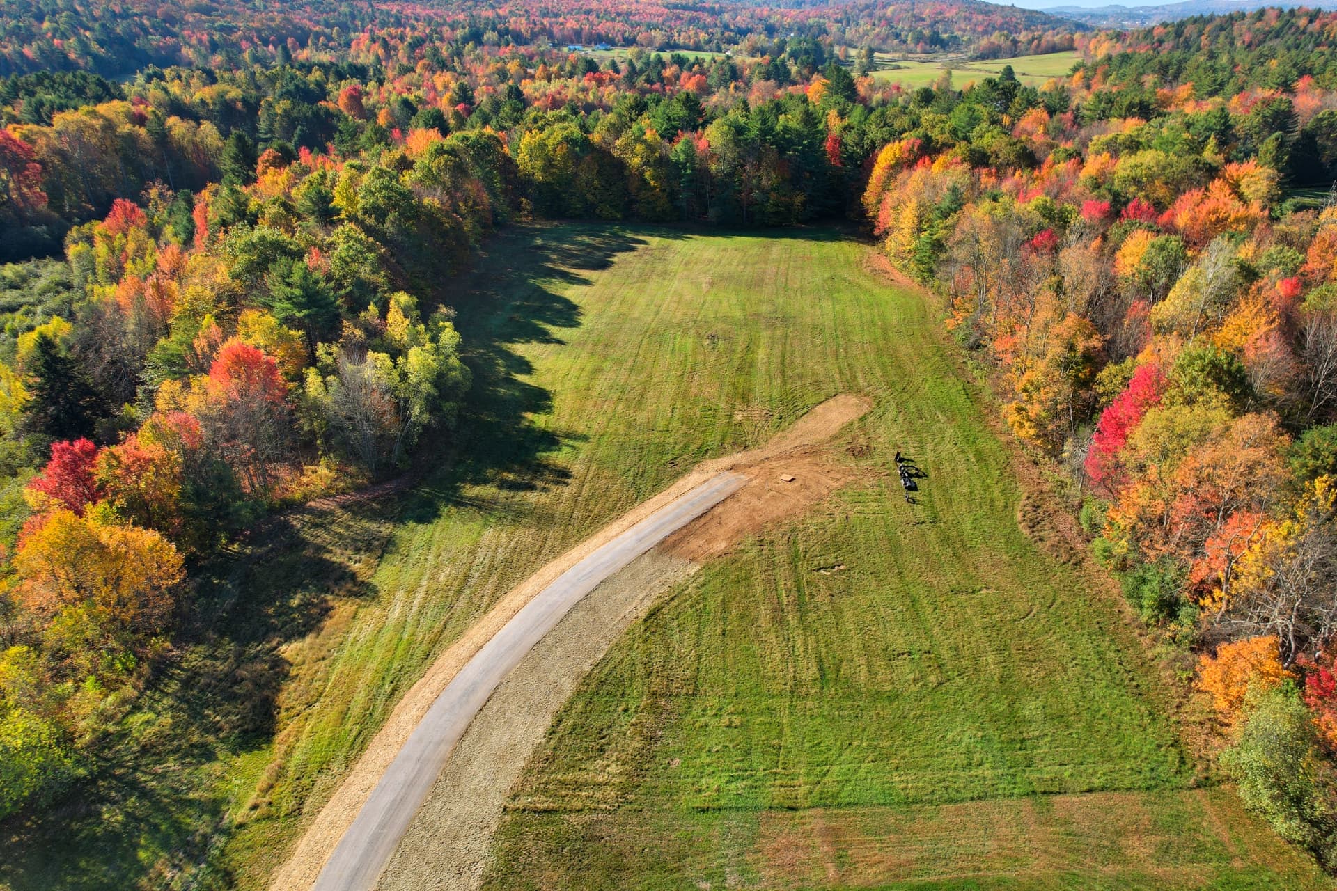 Top-down aerial of a new gravel road crossing a mowed lot ringed with red and gold foliage