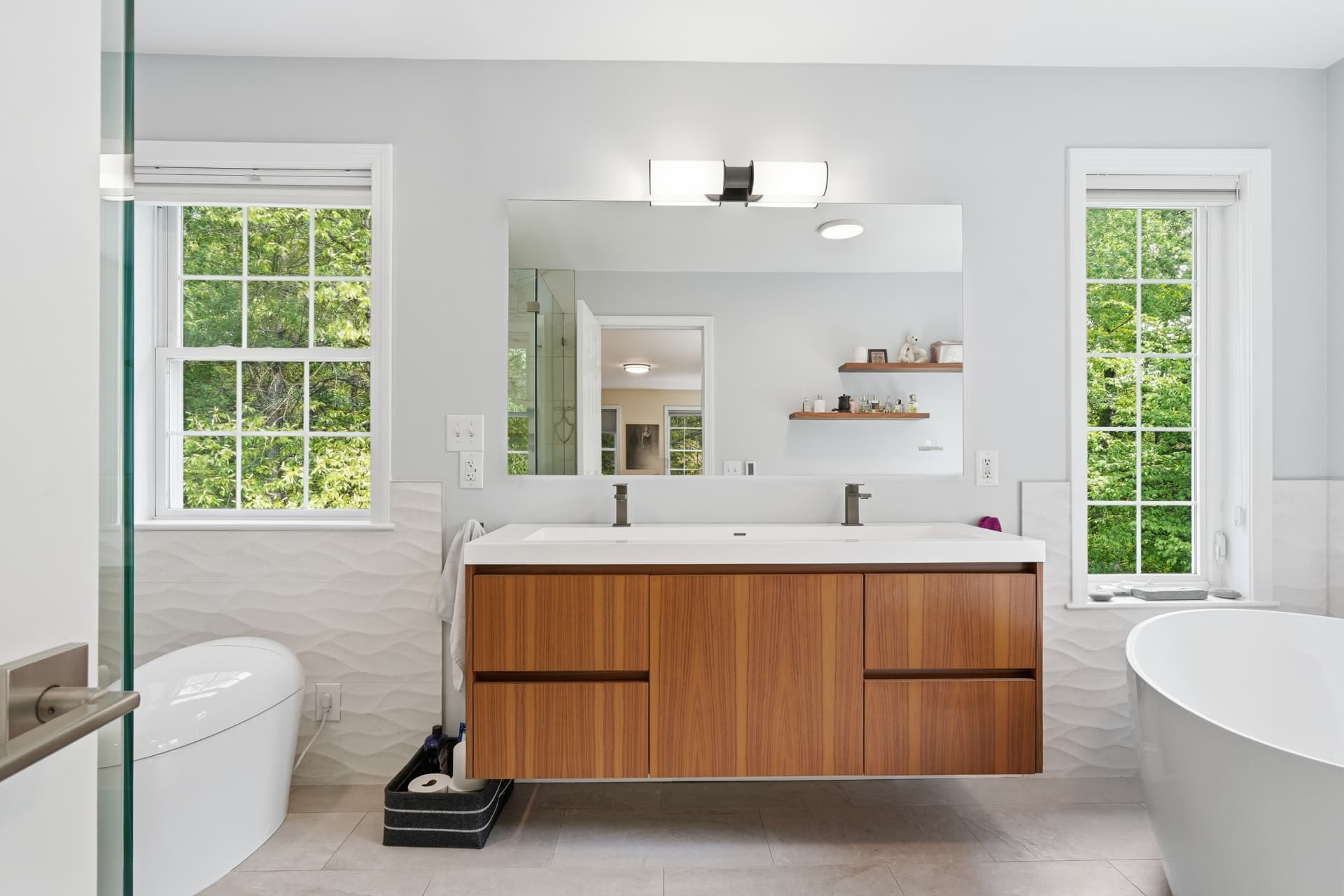 Bright bathroom with a wood floating double vanity, wide mirror, freestanding soaking tub and windows on both sides.