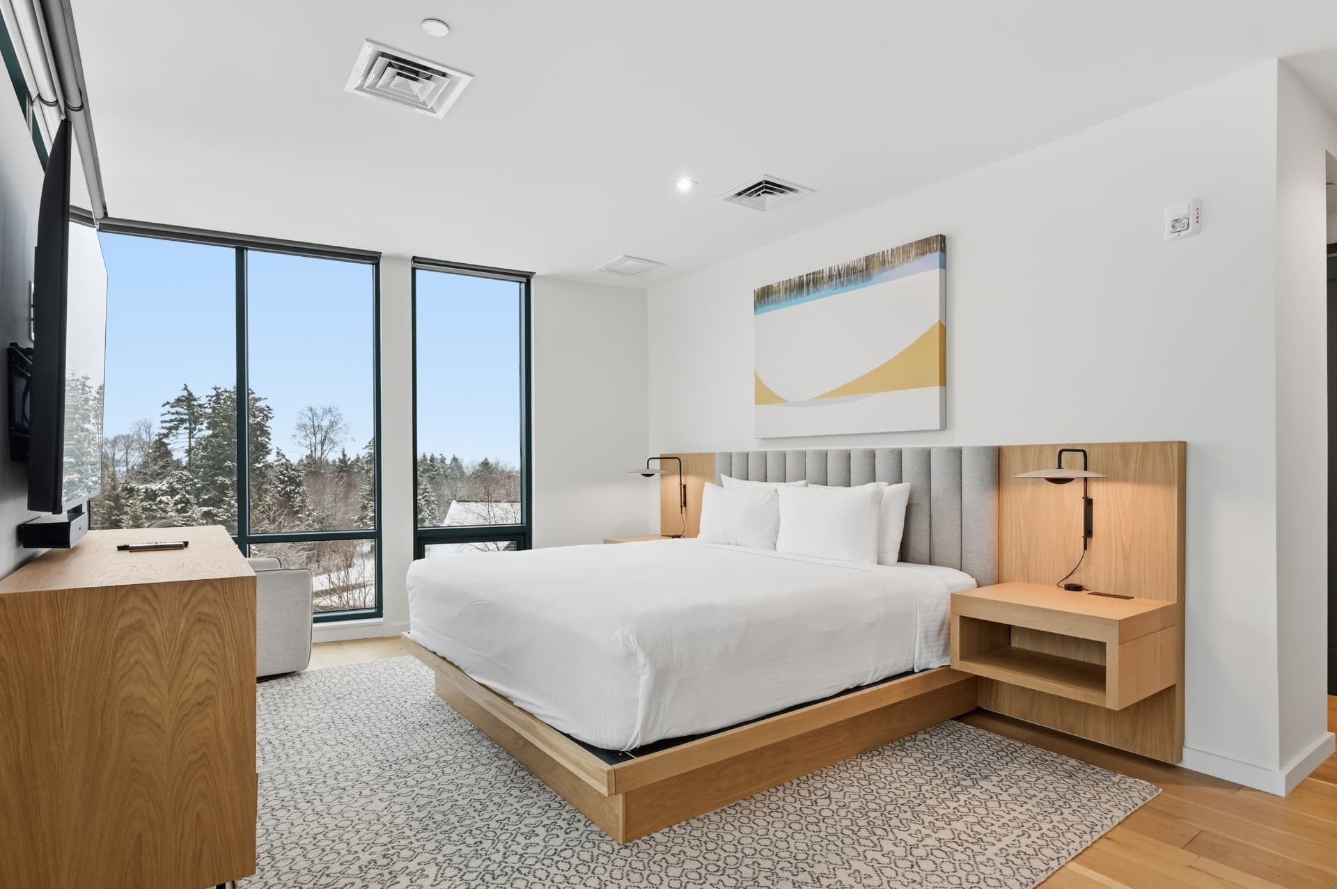 Modern hotel bedroom with a white bed, wood headboard and nightstands, large windows showing a snowy winter landscape.