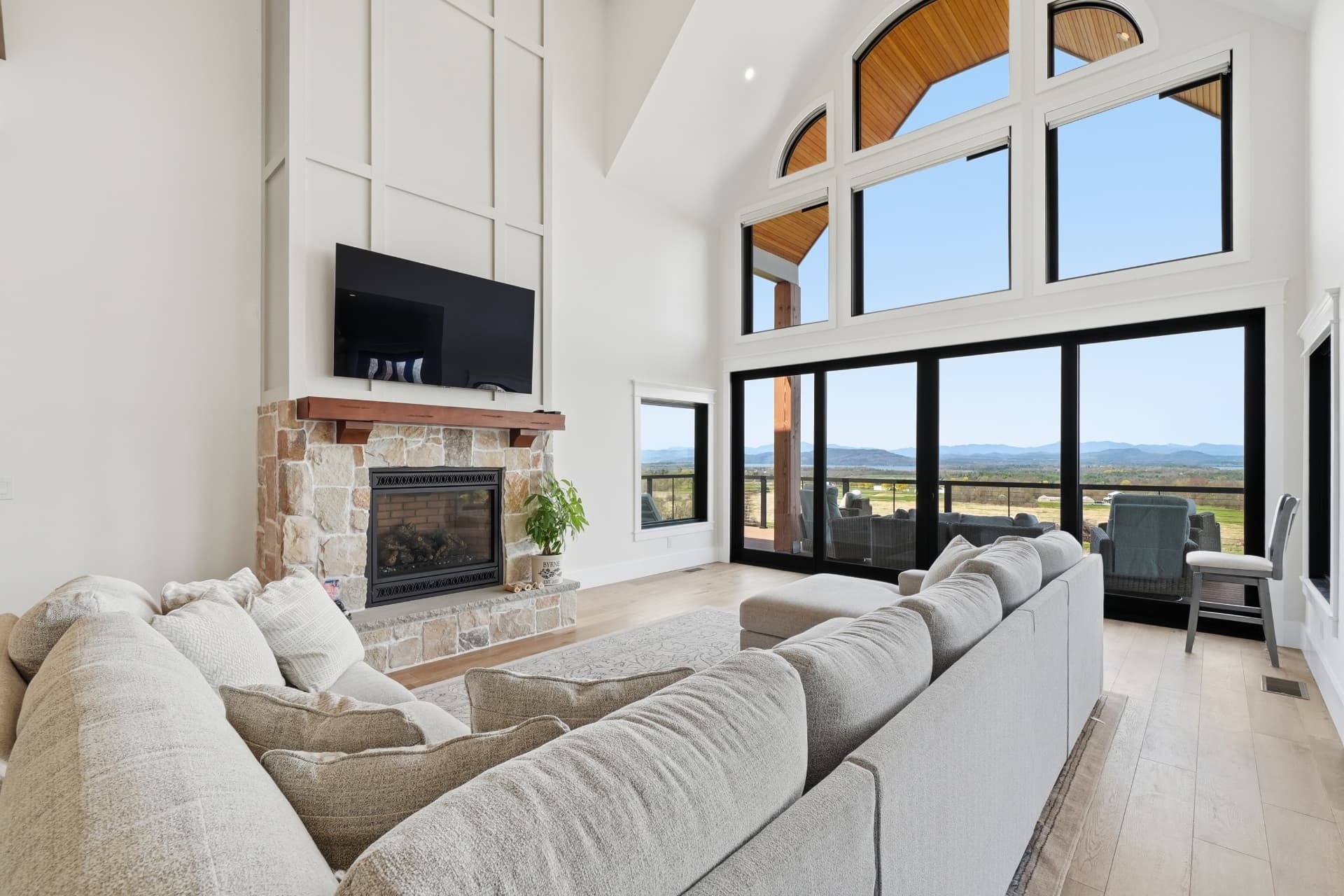 Two-story great room with a stone fireplace, wood mantel, large sectional sofa and tall windows framing a distant mountain view.