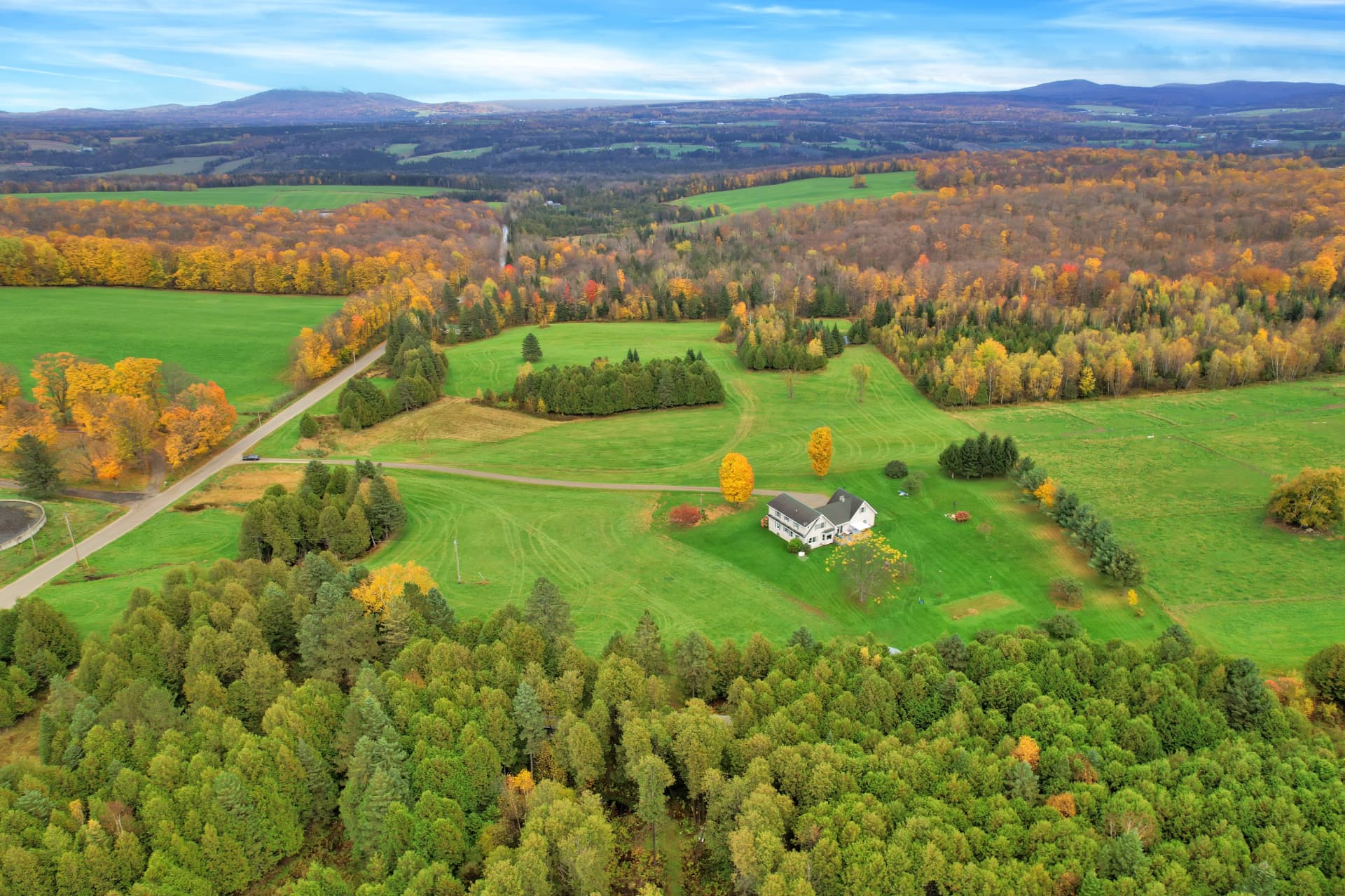 High aerial of a farmhouse on sweeping green acreage edged with autumn trees