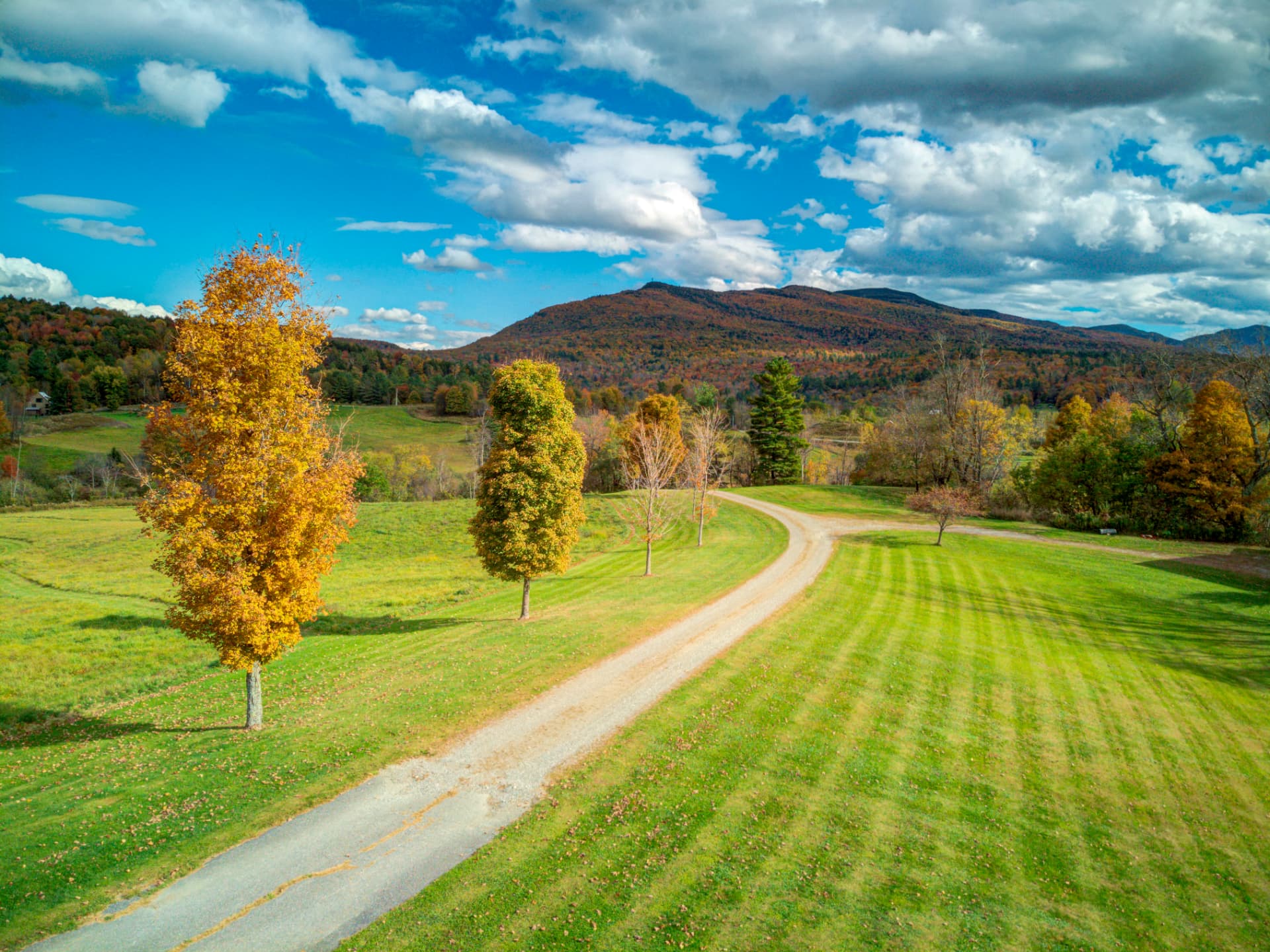 Aerial view of a gravel road through green pasture with fall foliage and Vermont mountains