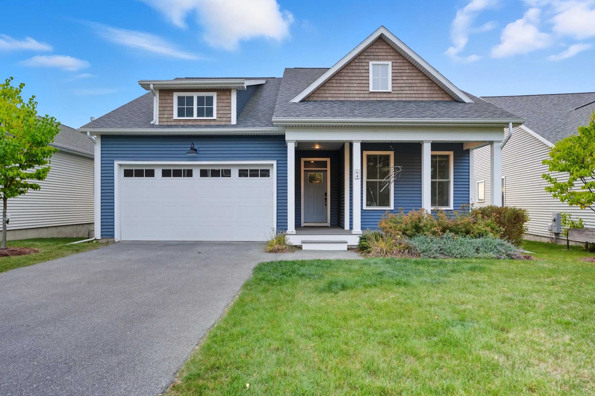 Blue-sided home with shingled dormers, a covered entry and a two-car garage, photographed from the paved driveway on a clear day.