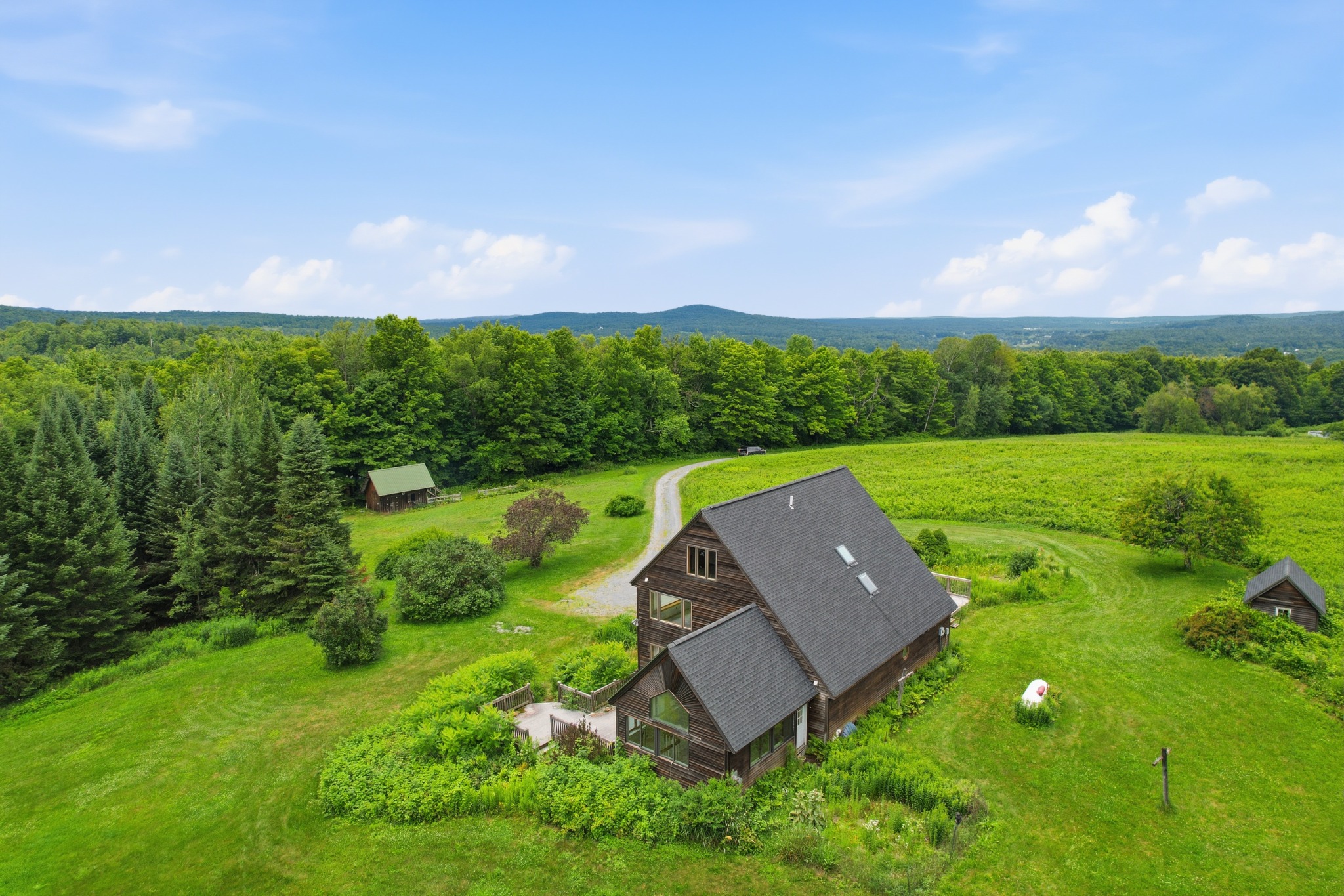 Aerial view of a dark-roofed home set into a green hillside meadow with a gravel drive and mature trees, mountains on the horizon.