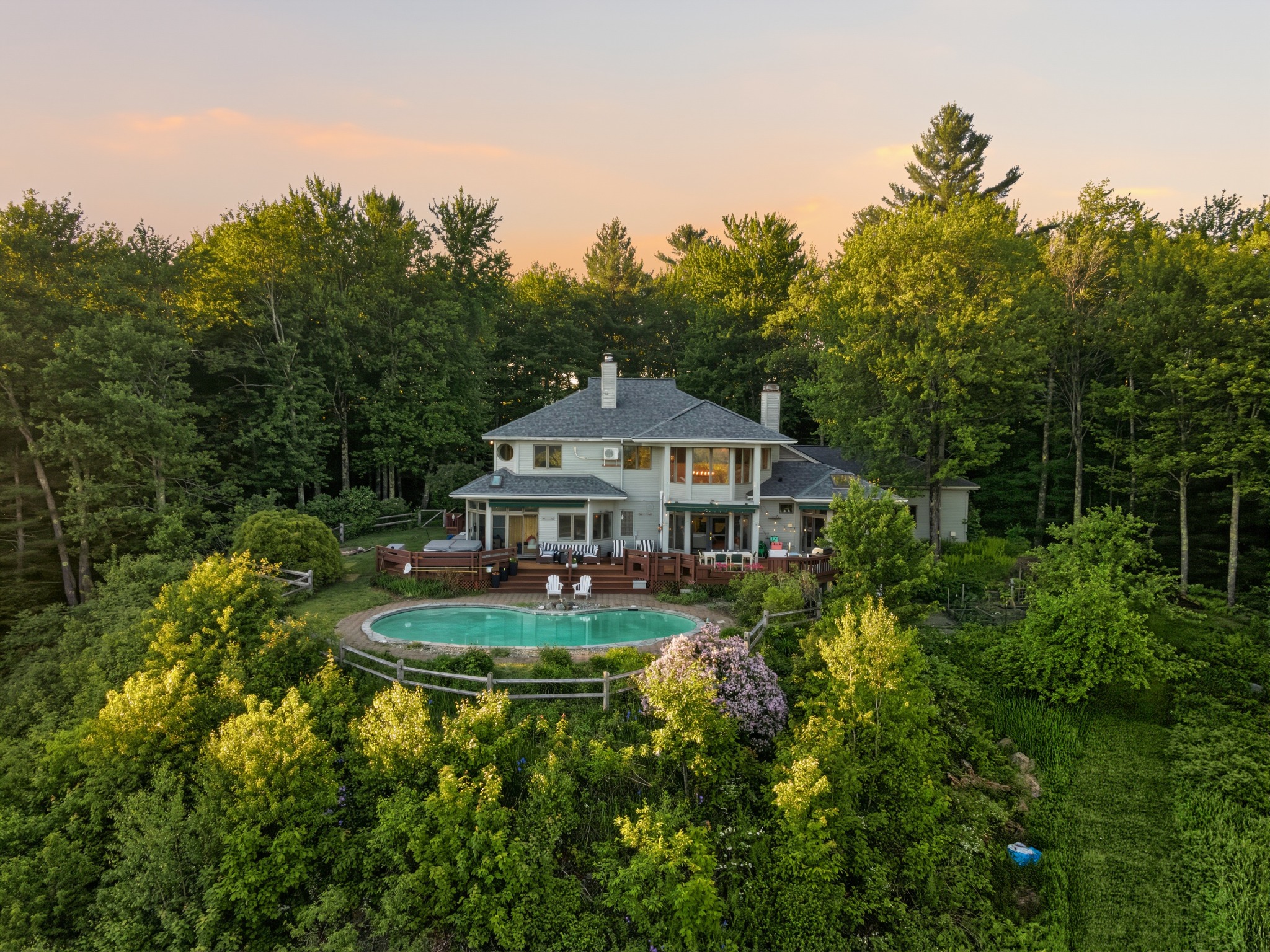 Aerial view of a two-story home with a wraparound porch and an oval swimming pool, surrounded by dense trees under a golden sunset sky.