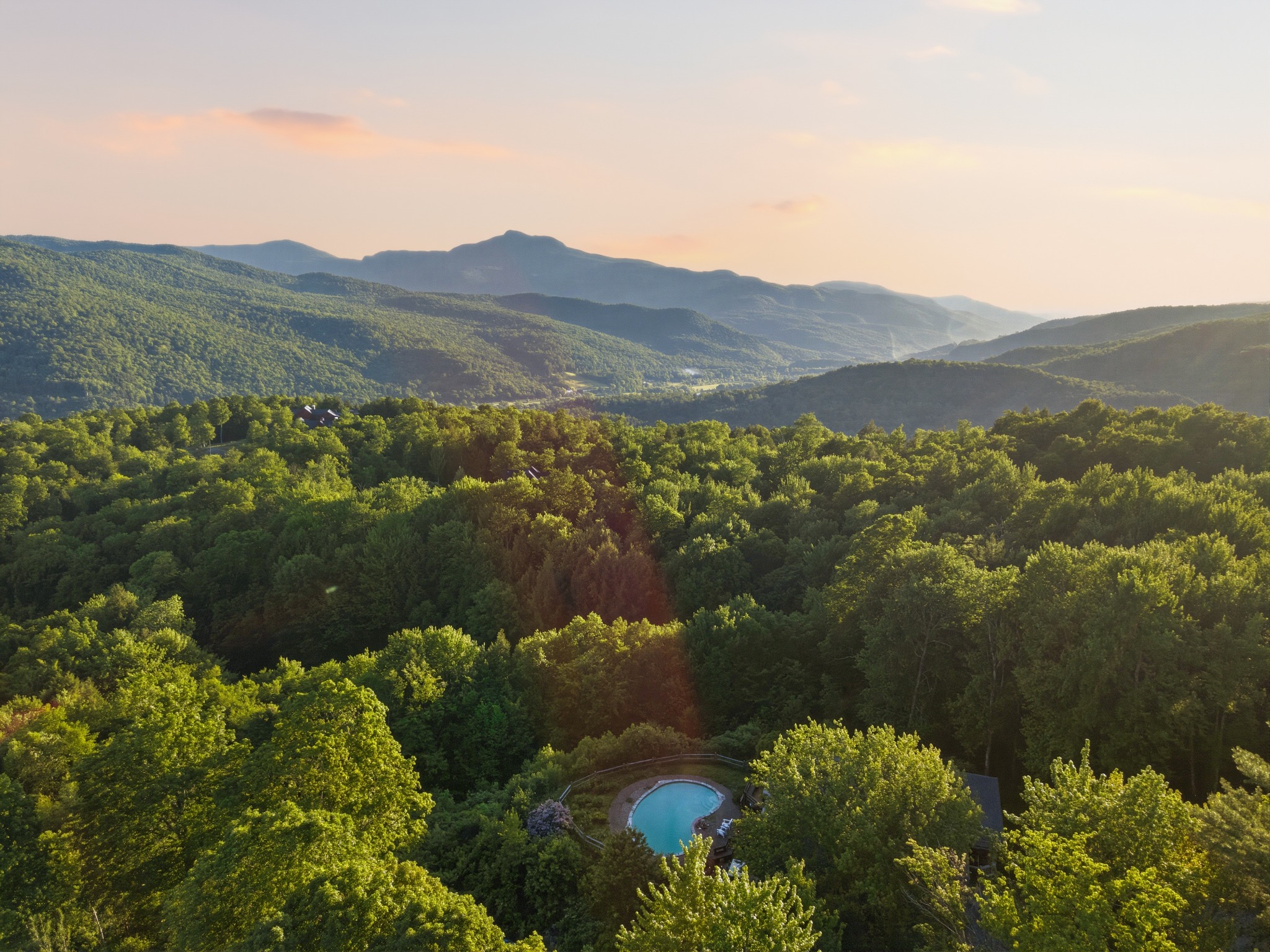 Aerial view over a forested valley at sunset with a distant mountain ridge and a small pool clearing among the trees.