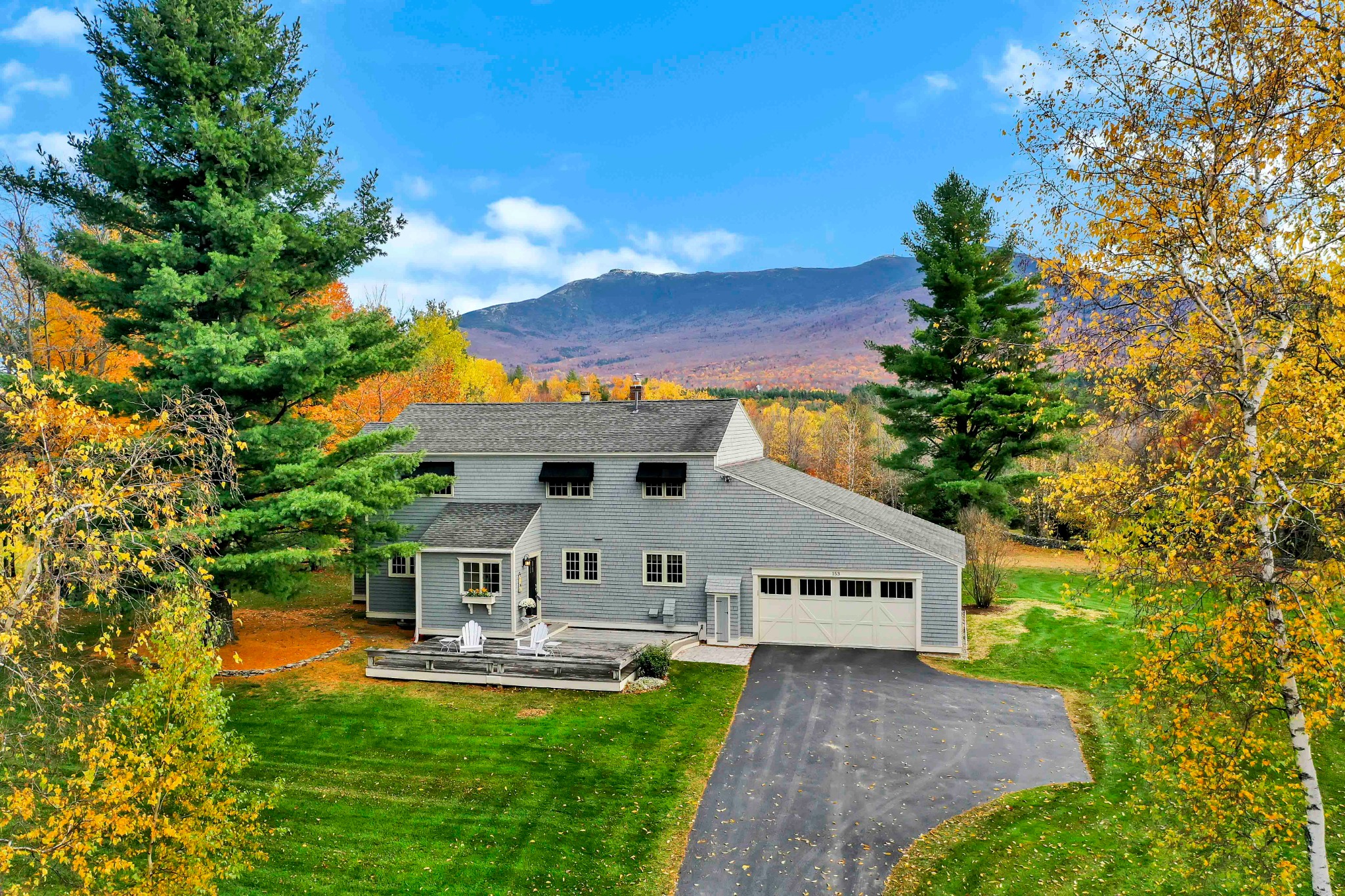 Aerial view of a gray home with an attached garage framed by yellow and orange autumn trees, with a broad mountain in the background.