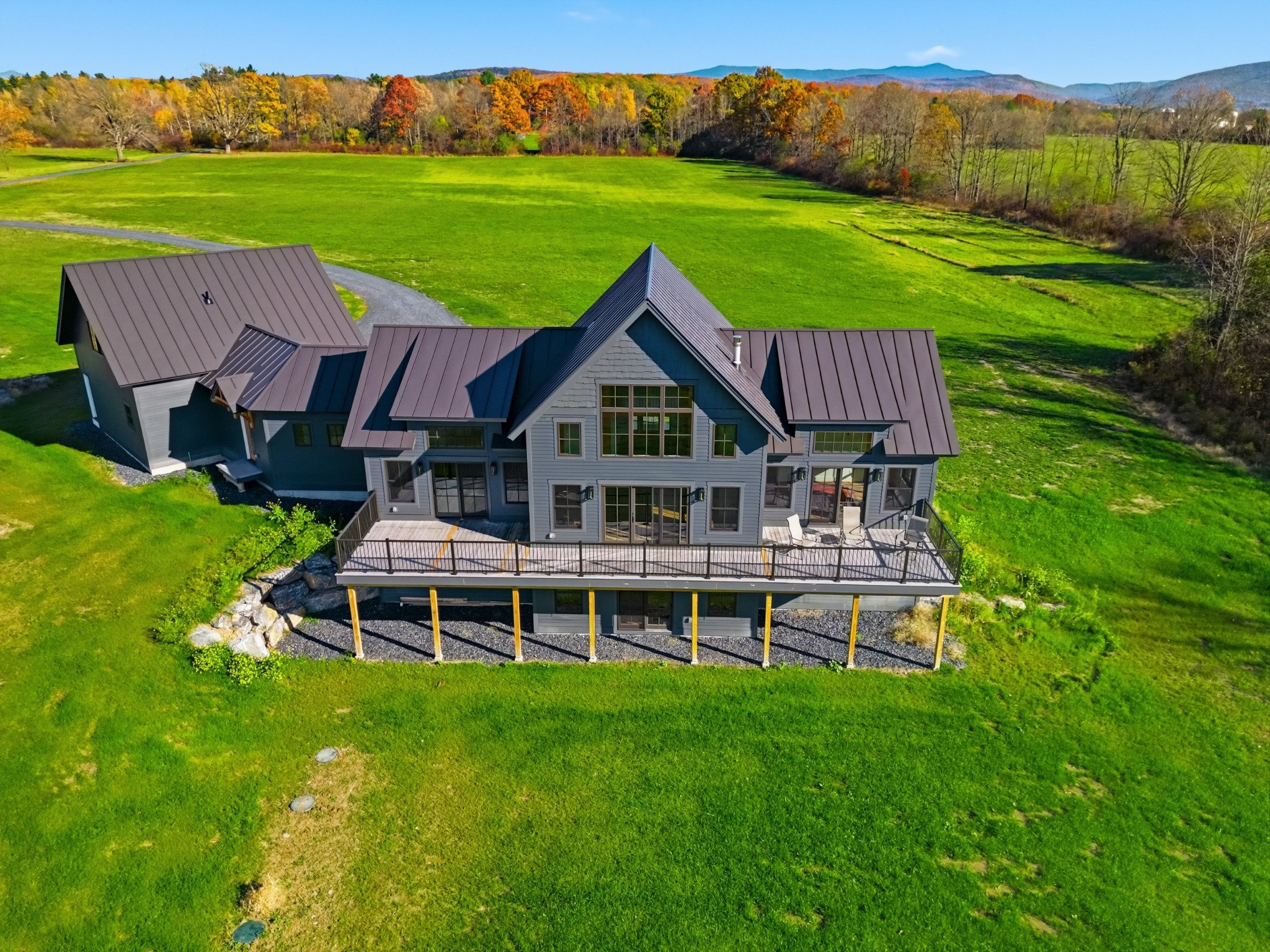 Aerial view of a modern gabled home with a metal roof and long deck sitting on a wide green field, with autumn woods and hills behind.