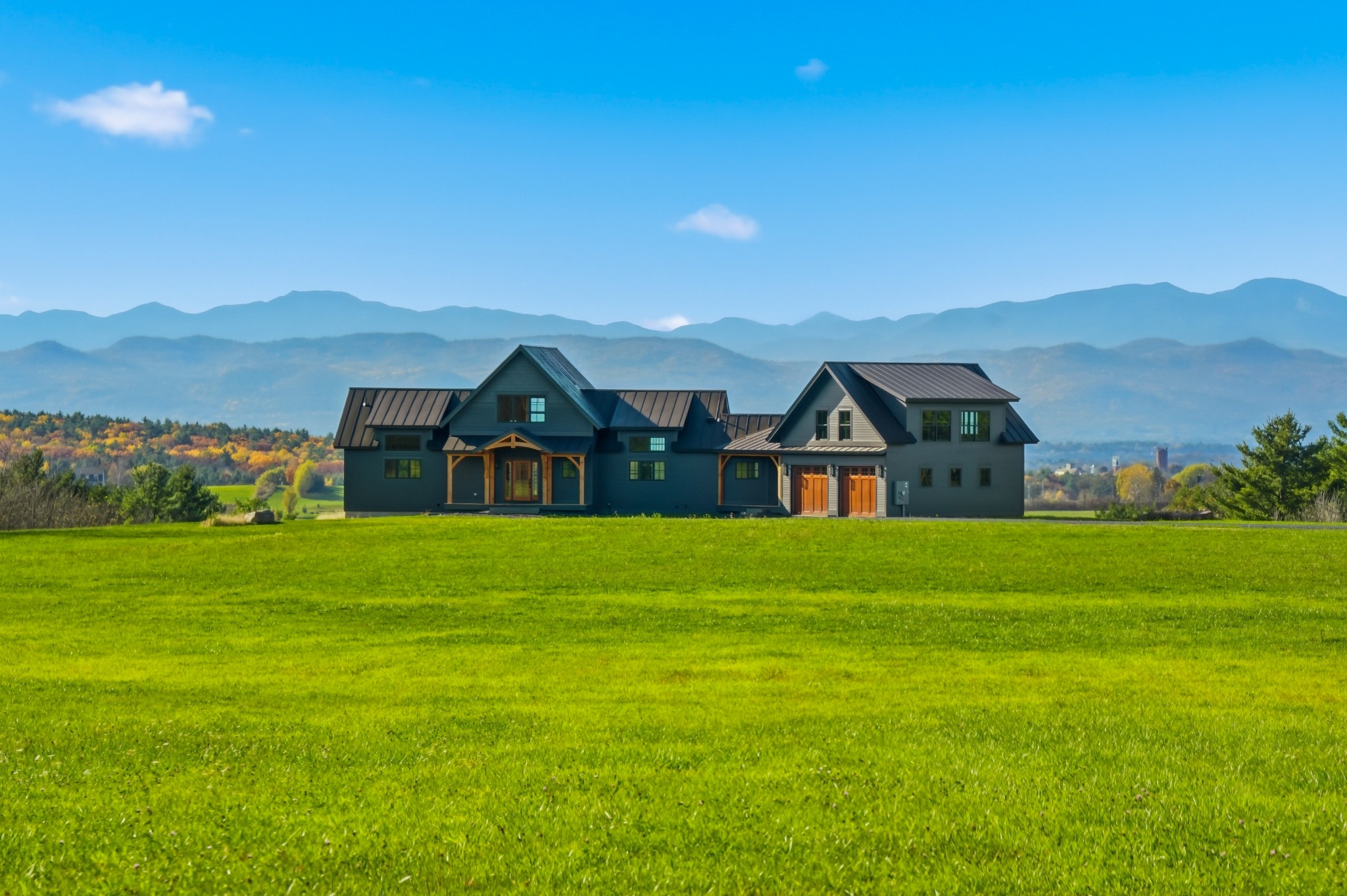 Dark modern farmhouse seen across a wide green field with a layered blue mountain range and scattered clouds behind it.