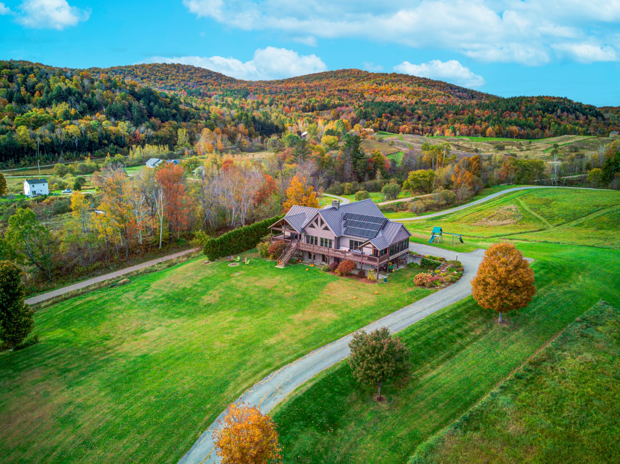Aerial view of a home with a curving driveway crossing rolling green pasture, with autumn-colored hills rising behind it.