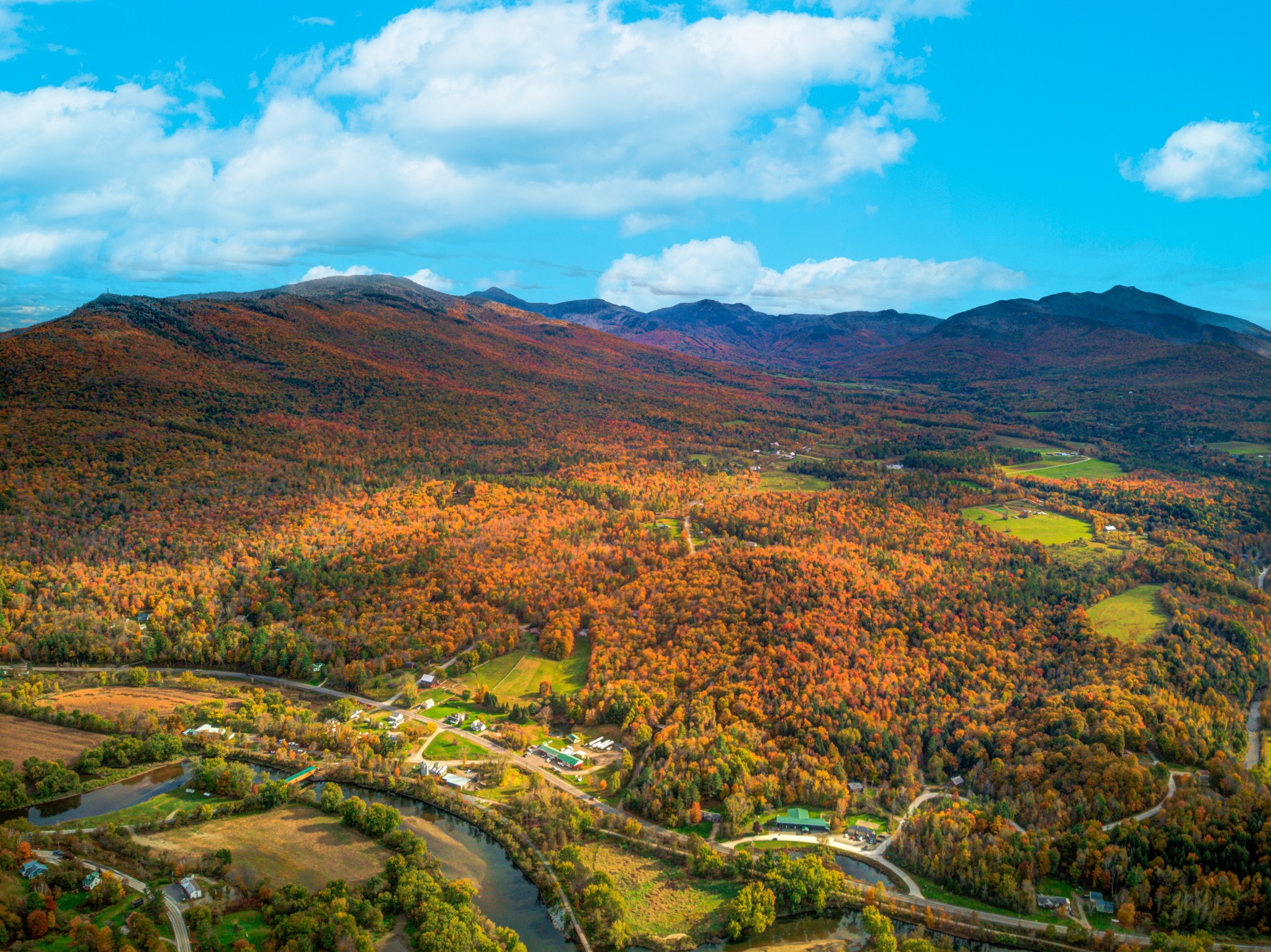 High aerial of a river valley in peak red and orange fall foliage with a road winding below distant blue mountains.
