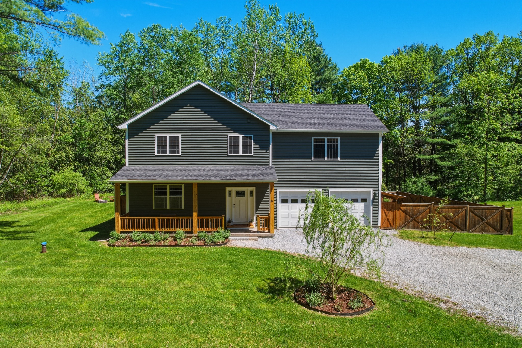 Gray two-story home with a covered front porch and attached garage, gravel driveway and a young tree in the front lawn.