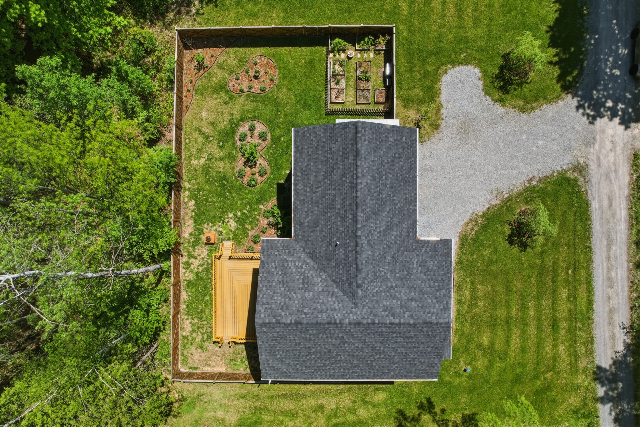 Straight-down aerial of a home's roofline showing the driveway, fenced yard and raised garden beds surrounded by trees.