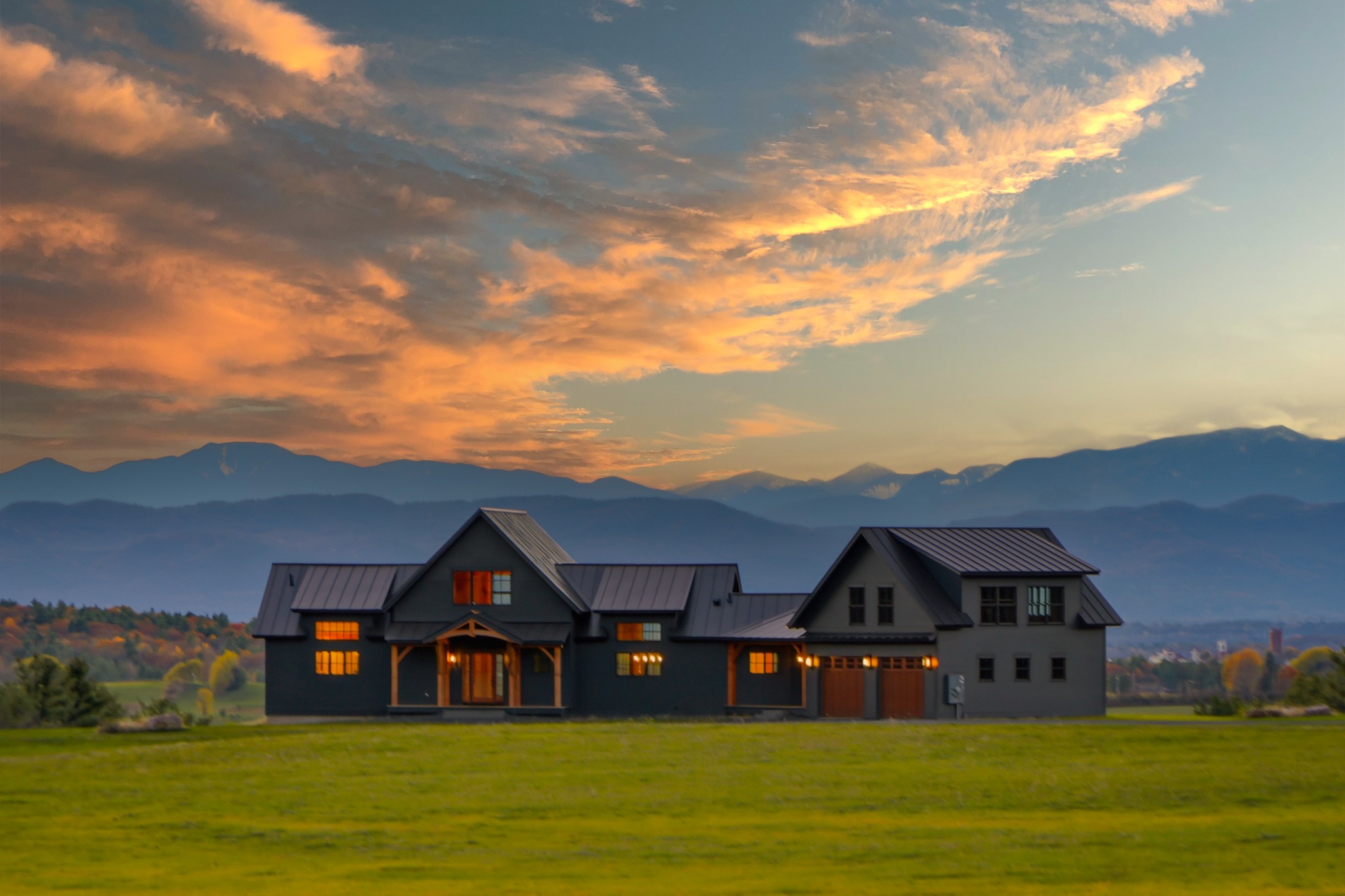 Modern farmhouse photographed at dusk with warm lit windows, a mountain ridge silhouette and orange streaked clouds behind it.