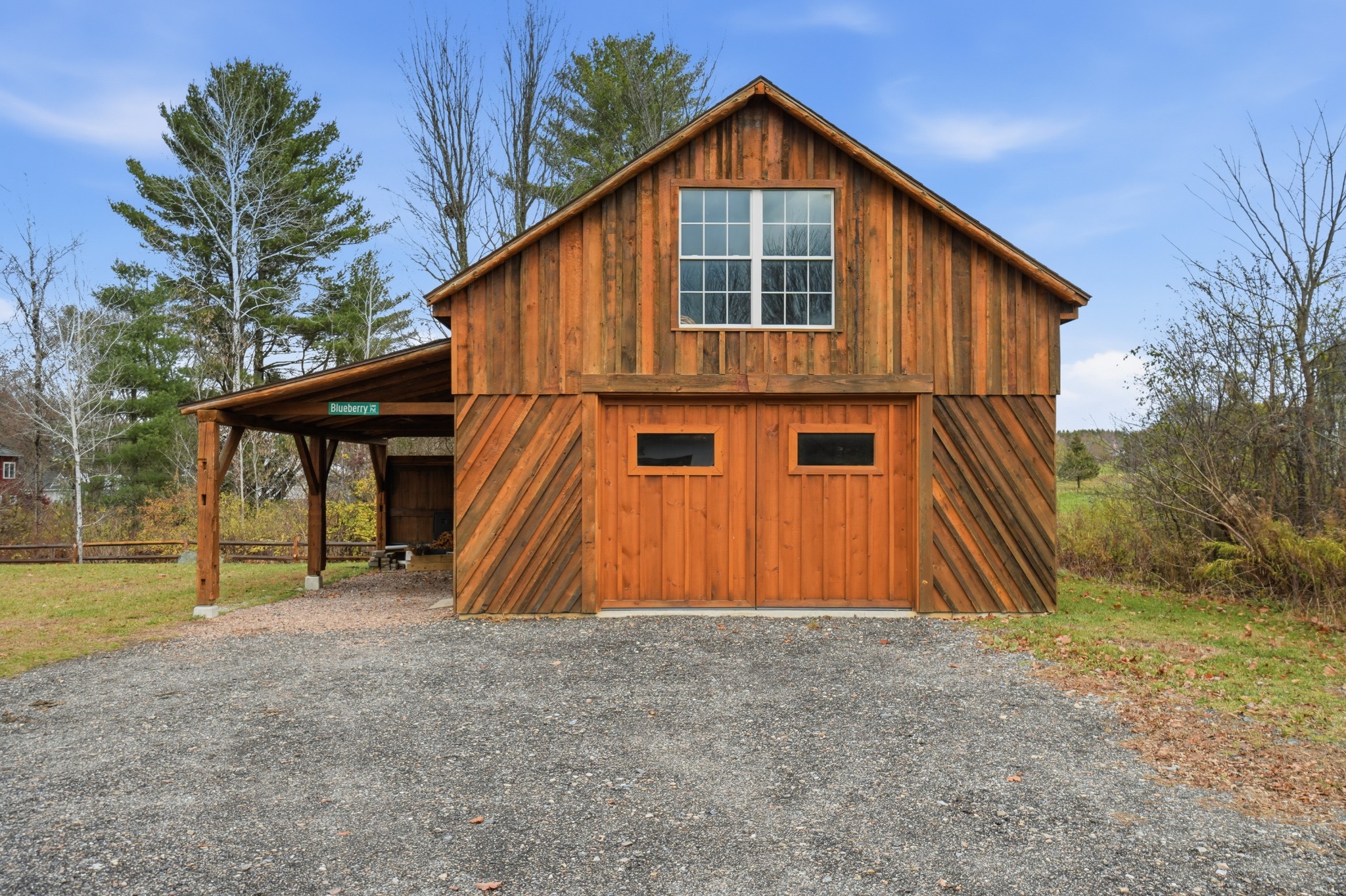 Wood-sided barn with a gable roof, upper window and covered side overhang, standing on a gravel pad under a blue sky.