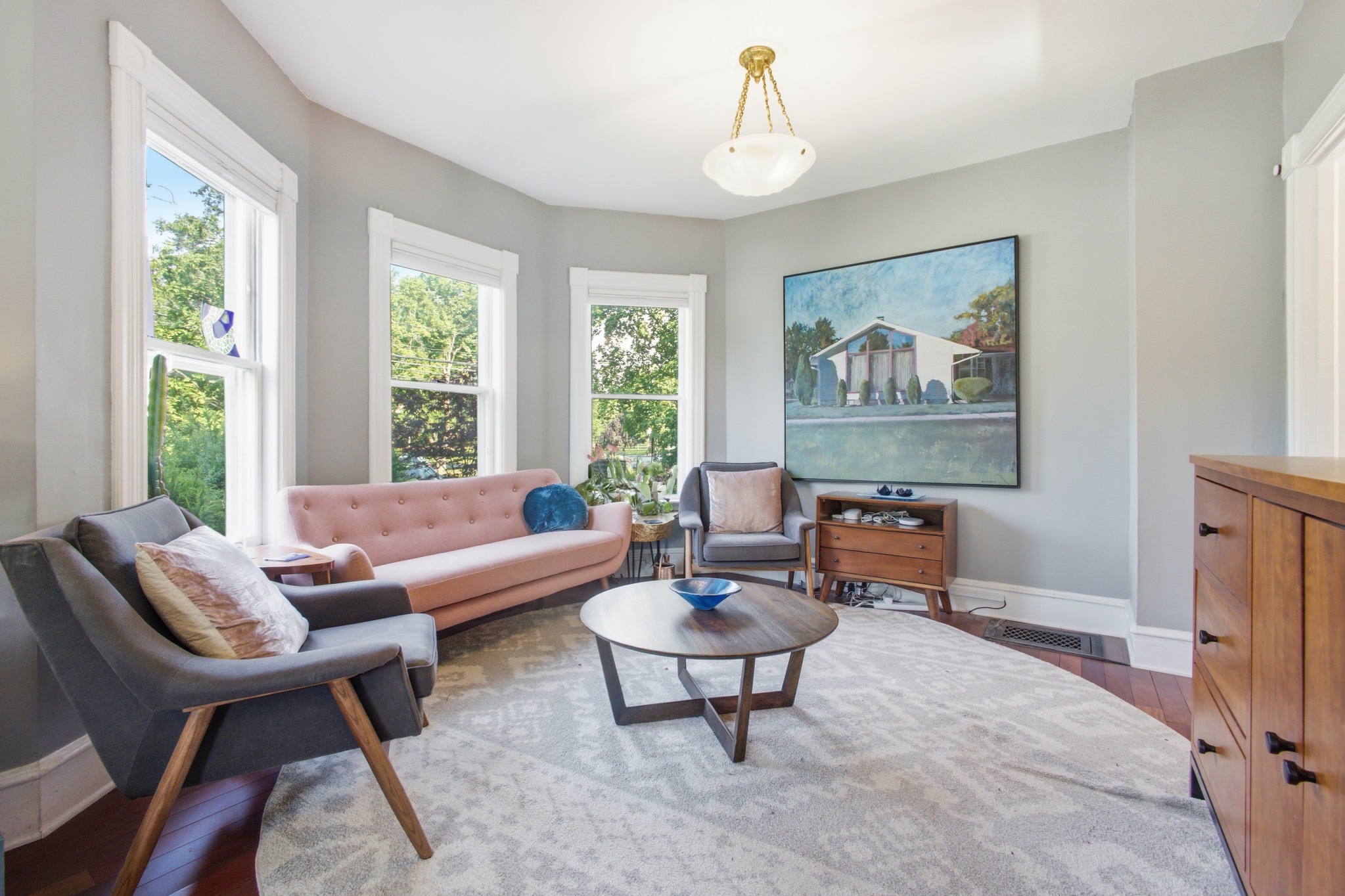 Living room with a pink sofa, gray armchairs, a round wood coffee table and bay windows looking into summer foliage.