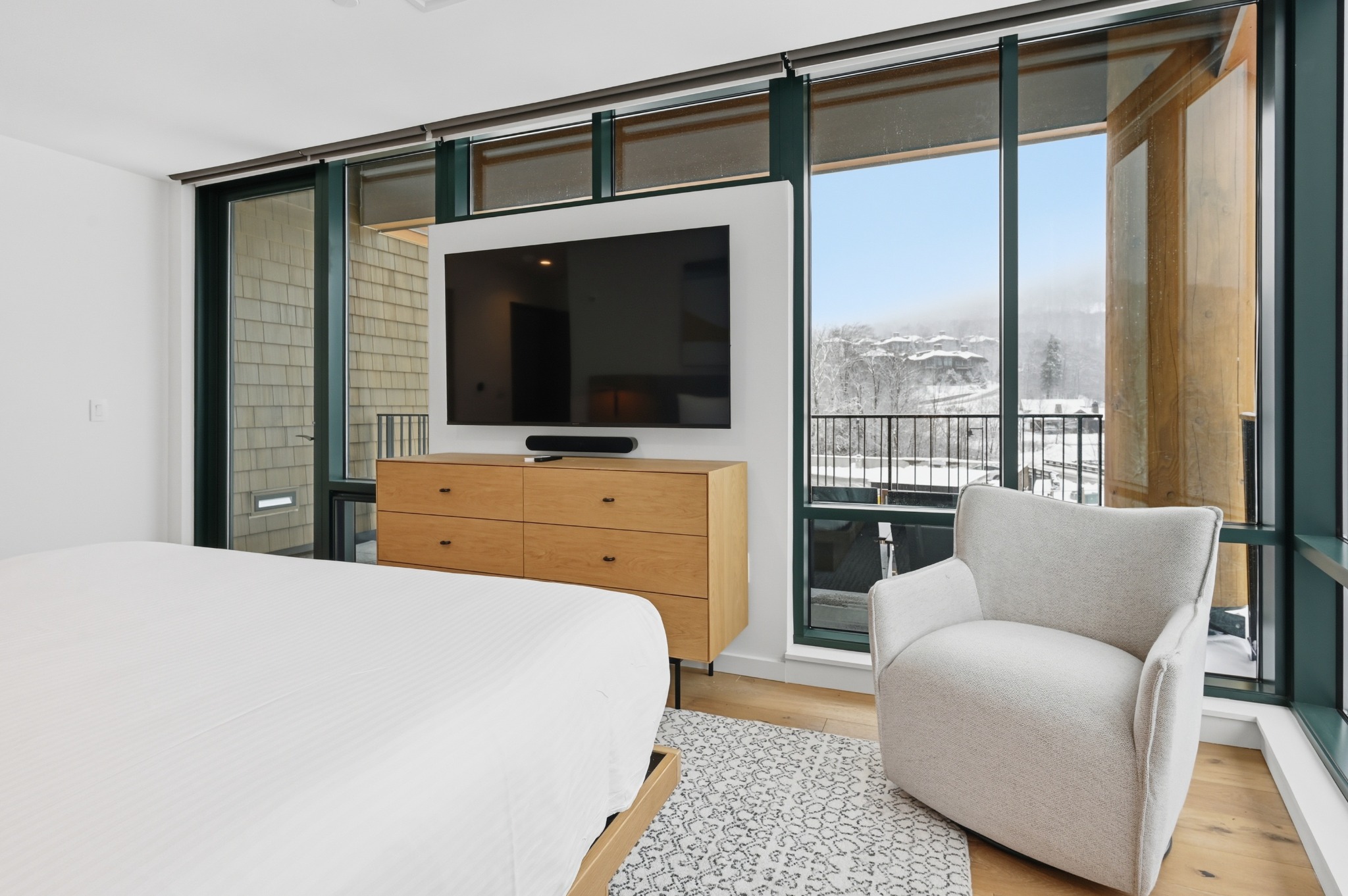 Bedroom with dark-framed glass walls opening to a balcony, a wall-mounted television, wood dresser and a white armchair.
