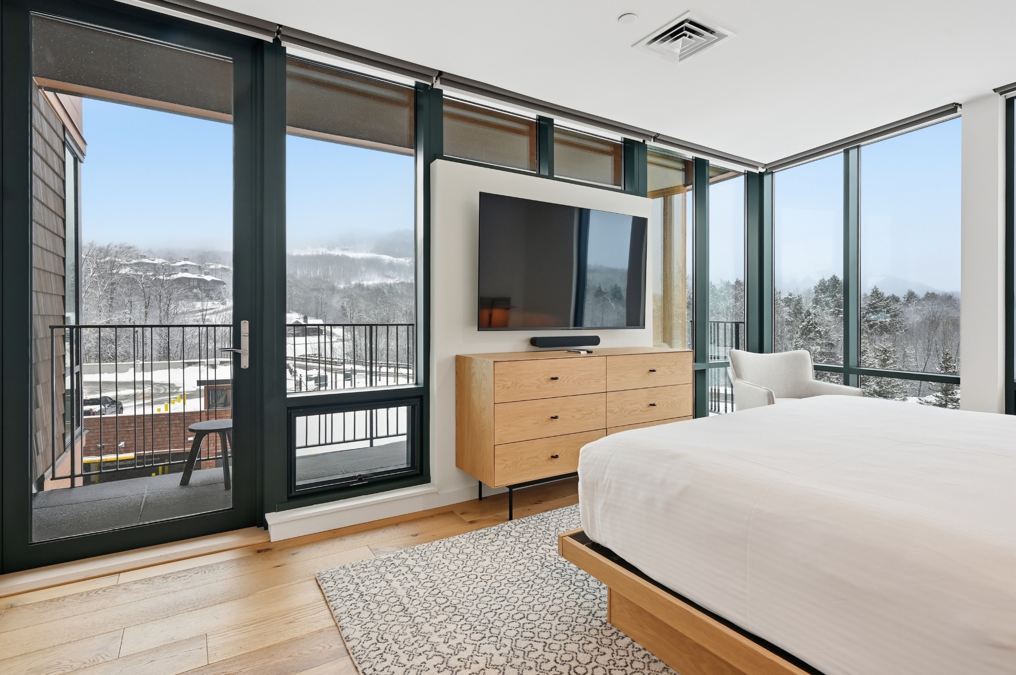 Wide view of a bedroom wrapped in dark-framed windows on two sides, with a platform bed, dresser and patterned area rug.