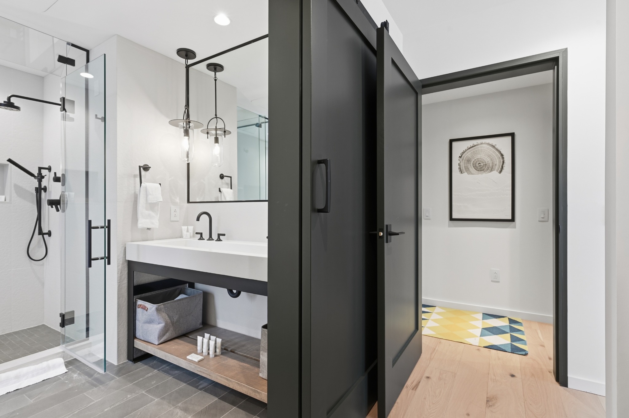 Bathroom with a glass walk-in shower, floating vanity and round mirror, seen through an open black-framed door.