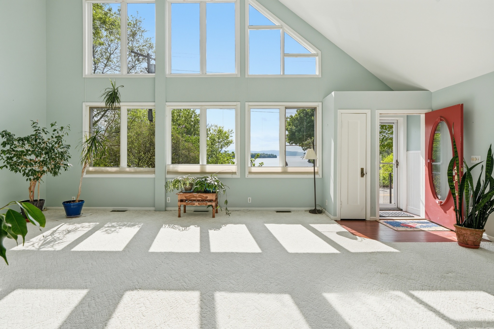 Two-story great room with a bank of tall windows, sage green walls, potted plants and a red front door, sunlight across the carpet.