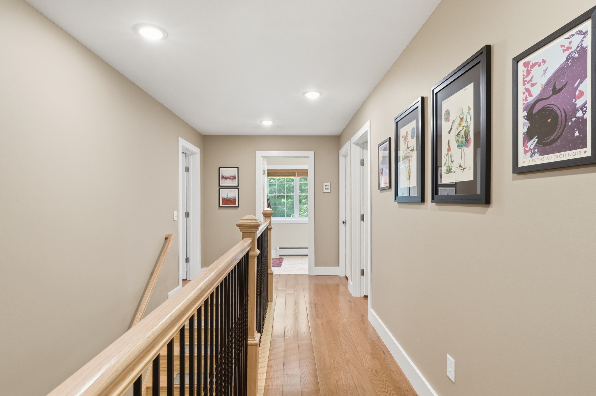 Upstairs hallway with wood flooring, a stair railing and a wall of framed artwork leading toward a bright window.