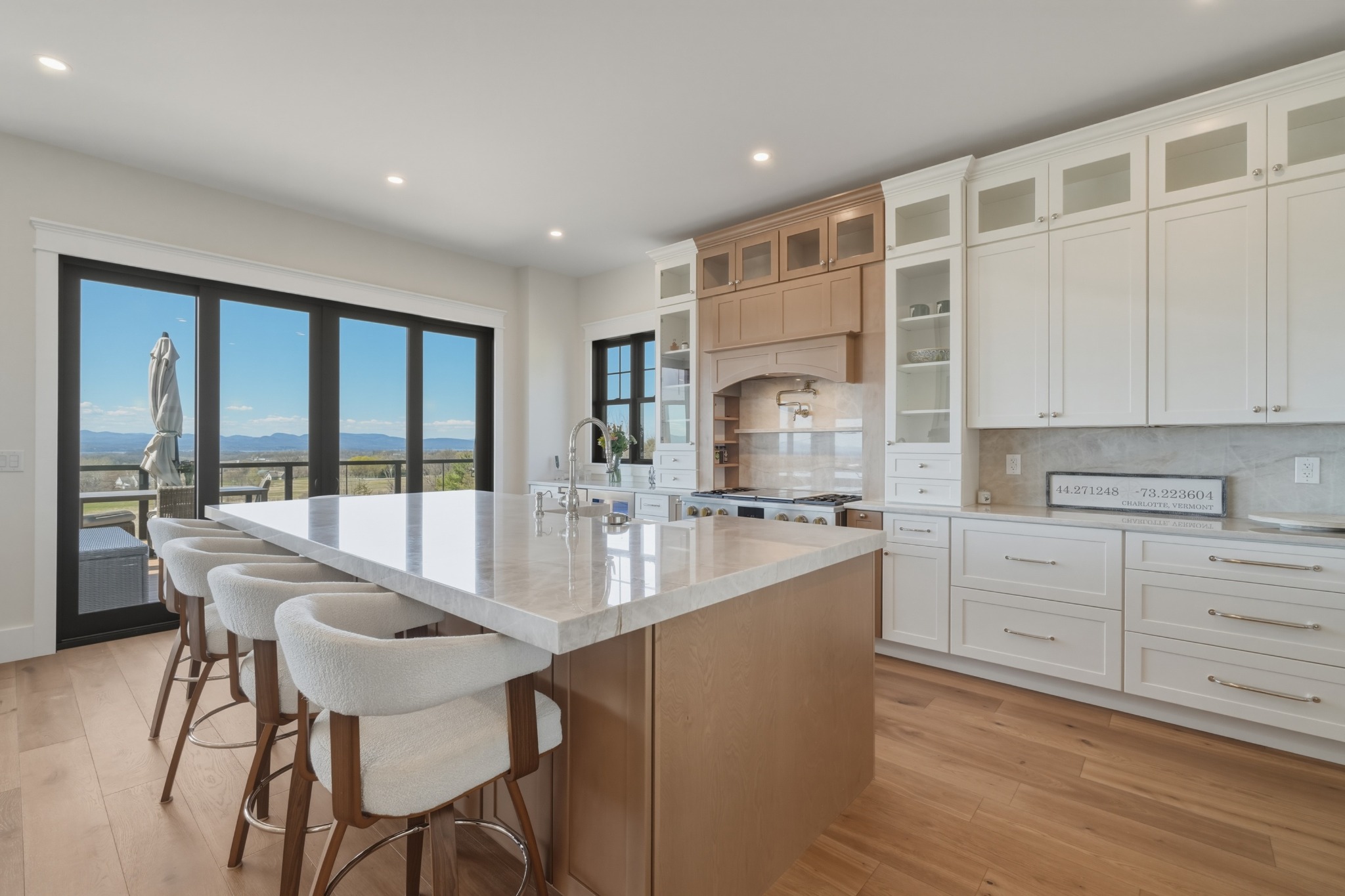 White kitchen with a long quartz island, upholstered stools, glass-front cabinets and wide windows framing a distant mountain view.