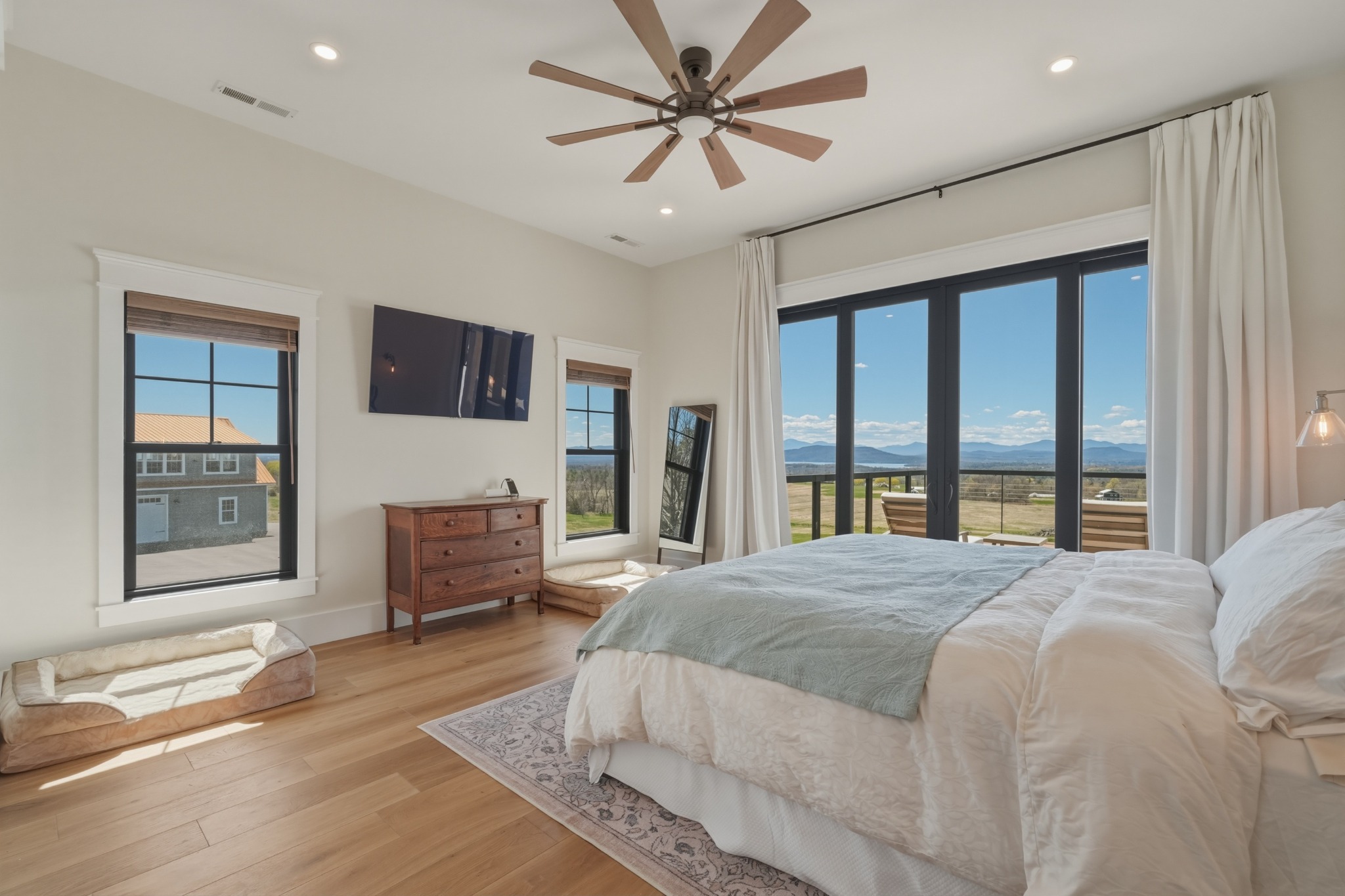 Bedroom with a large ceiling fan, white bedding and tall windows showing a distant mountain horizon beyond the yard.